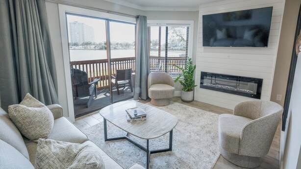 A modern living room with a sofa, armchair, coffee table, TV, fireplace, and a view through the glass door. - 112 Arrowhead – Unique Lakefront 2BR at Kingston (Myrtle Beach)