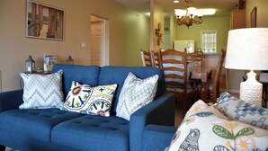 A living room with a blue sofa, patterned cushions, a dining area with wooden chairs, and a chandelier. - New 2026 Listing! Pond View Condo in Five Points (Lewes)