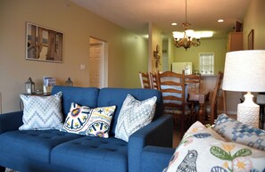 A living room with a blue sofa, patterned cushions, a dining area with wooden chairs, and a chandelier. - New 2026 Listing! Pond View Condo in Five Points (Lewes)