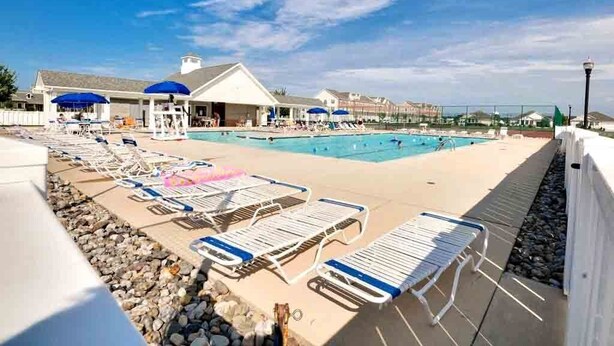 A pool area with lounge chairs and umbrellas, a building with a sloped roof, and a clear sky. - New 2026 Listing! Pond View Condo in Five Points (Lewes)