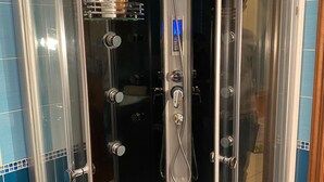 A glass-enclosed shower stall with a control panel, a shelf with toiletries, and a tiled floor. - Appartamento confortevole con doccia idromassaggio (Torino)