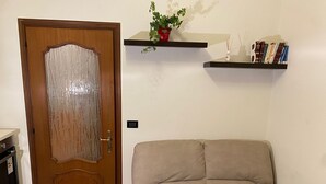 A room with a wooden door, a beige sofa, a small plant on a shelf, and a book collection on another shelf. - Appartamento confortevole con doccia idromassaggio (Torino)
