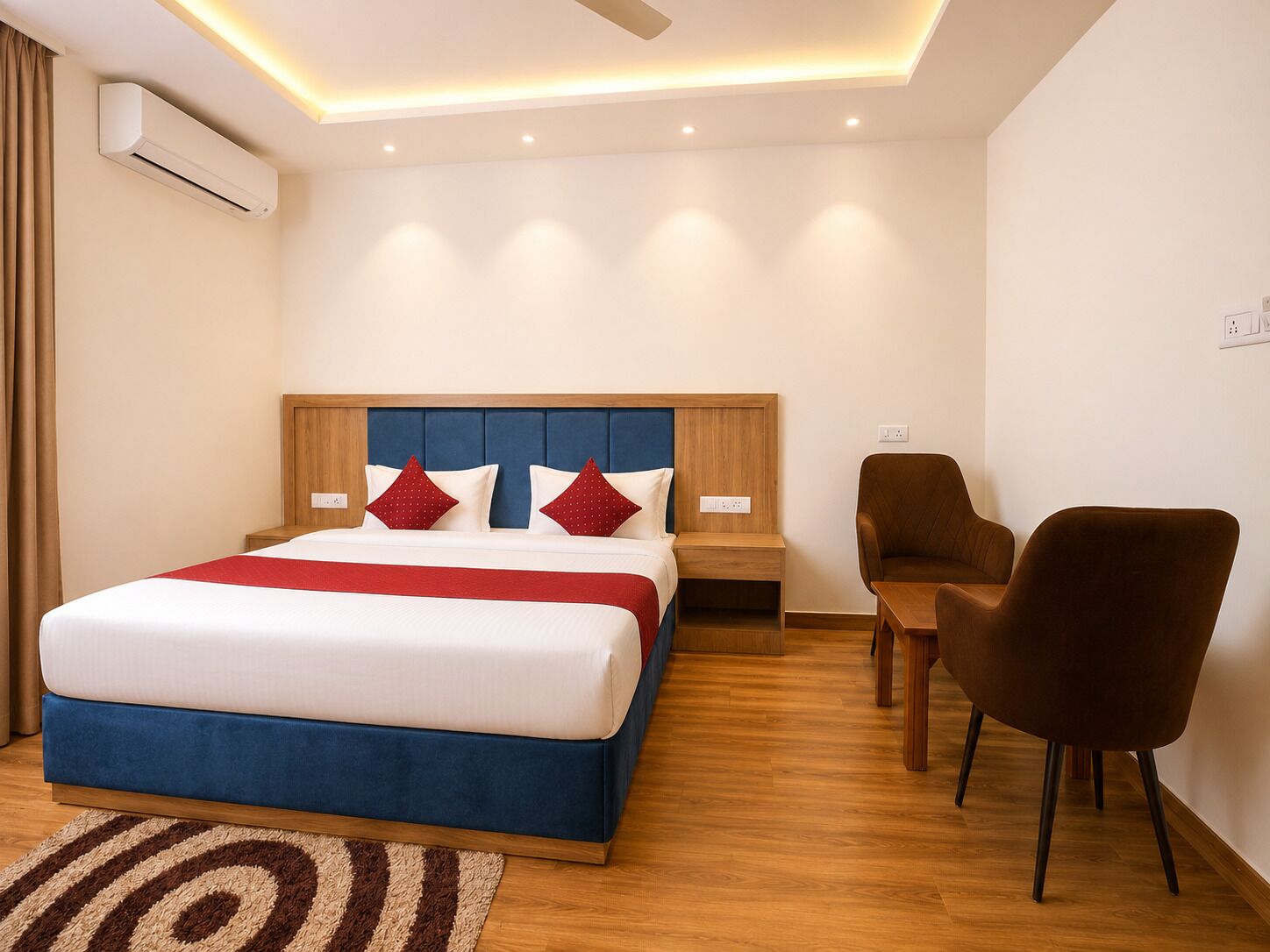 A hotel room with a bed, bedside tables, a desk with chairs, an air conditioning unit, and a ceiling fan.