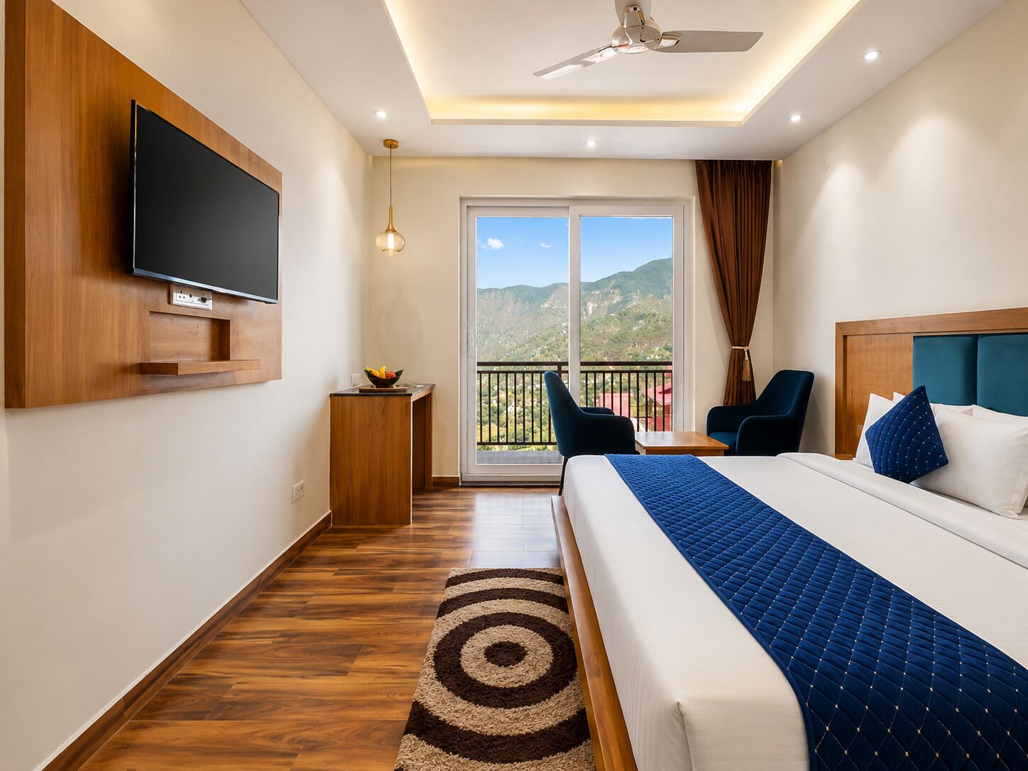 Camera d'albergo con un letto, una televisione a parete, un tavolino in legno, un balcone con sedie e vista sulle montagne.