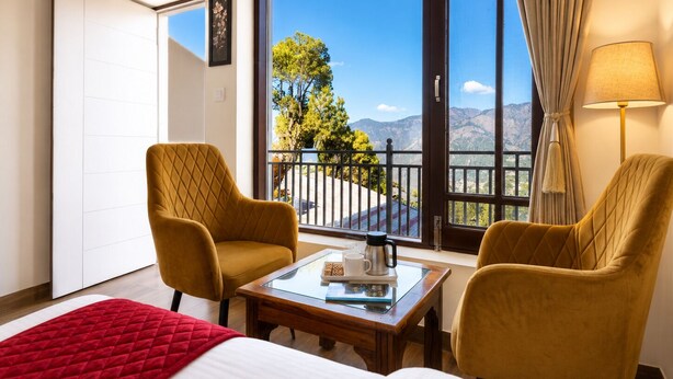 Ein Hotelzimmer mit einem Bett, zwei Sesseln, einem kleinen Tisch, einem Balkon mit Blick auf Berge und Bäume sowie einem Vorhangfenster.