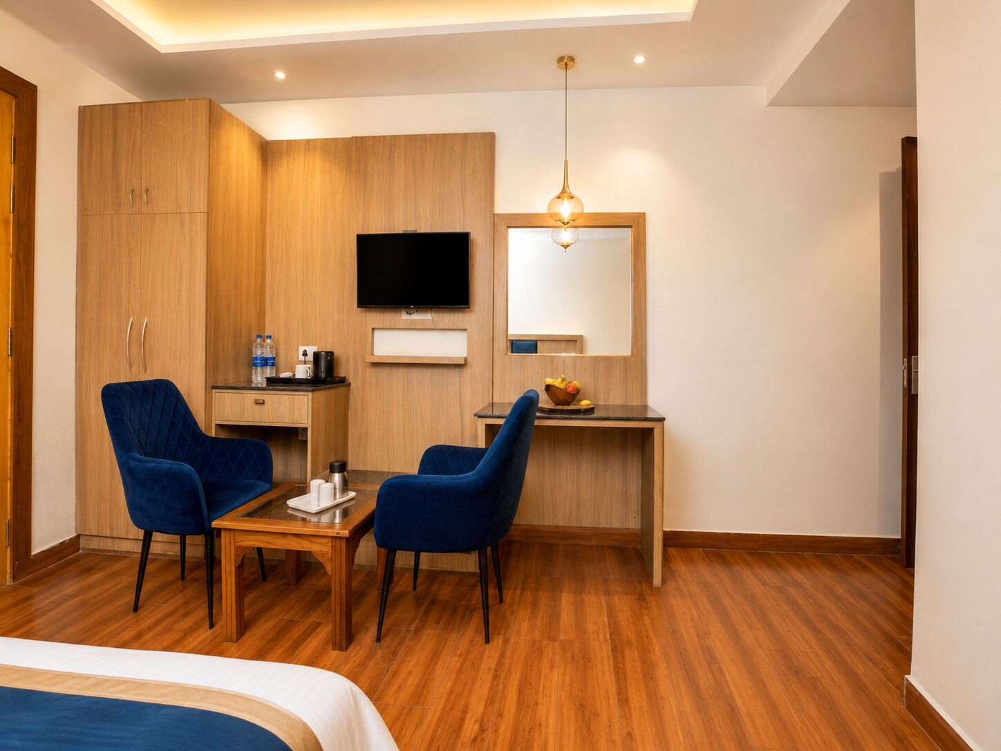 Quarto de hotel com cama, duas poltronas azuis, uma secretária de madeira com televisão de tela plana, um espelho e um candeeiro de teto.
