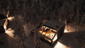 Een besneeuwde hut met een verlicht interieur, omringd door bomen.