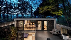 Una casa moderna con fachada de vidrio, un patio con mobiliario y vistas al bosque circundante.