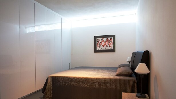 Un dormitorio con cama, mesita de noche, lámpara, una obra de arte enmarcada en la pared y un armario.