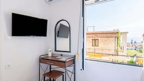 A room with a desk, a flat-screen TV, a mirror, and a window with a view of buildings and a balcony. - Gavriil Apartments Corfu - Courtyard Retreat (Corfu)
