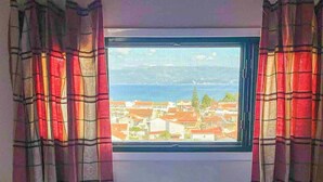 Ein Bett mit weißen Leinen und Handtüchern, ein Fenster mit Blick auf Gebäude und ein Gewässer, sowie karierte Vorhänge.