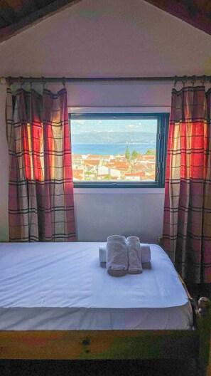 Una cama con ropa de cama y toallas blancas, una ventana con vistas a edificios y un cuerpo de agua, y cortinas de cuadros.