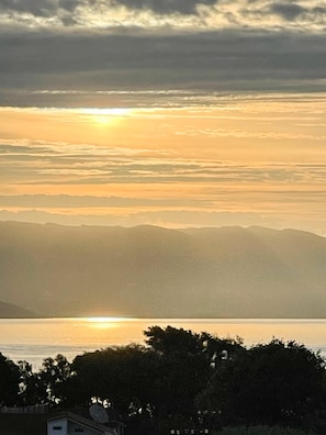 Un coucher de soleil sur une étendue d’eau, avec des montagnes en arrière-plan et des arbres au premier plan.