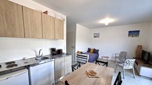A compact kitchen with a sink, stove, and coffee maker, a dining area with a table and chairs, and a bed with pillows. - The "Le Cosy" Apartment – Shared Pool (La Tranche-sur-Mer)