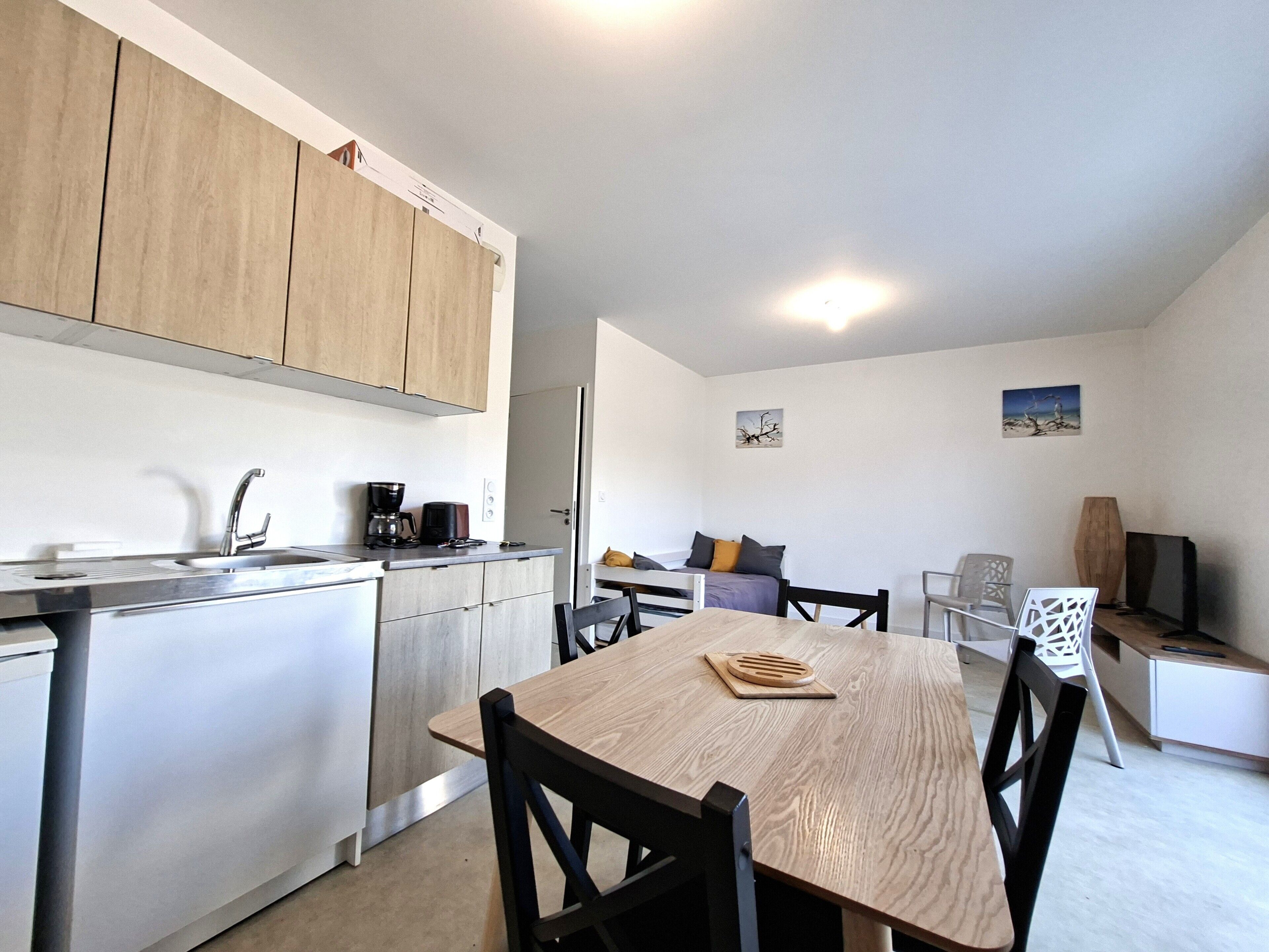 Cocina compacta con lavamanos, cafetera y armarios. Hay un comedor con mesa y sillas, y un televisor en una esquina.