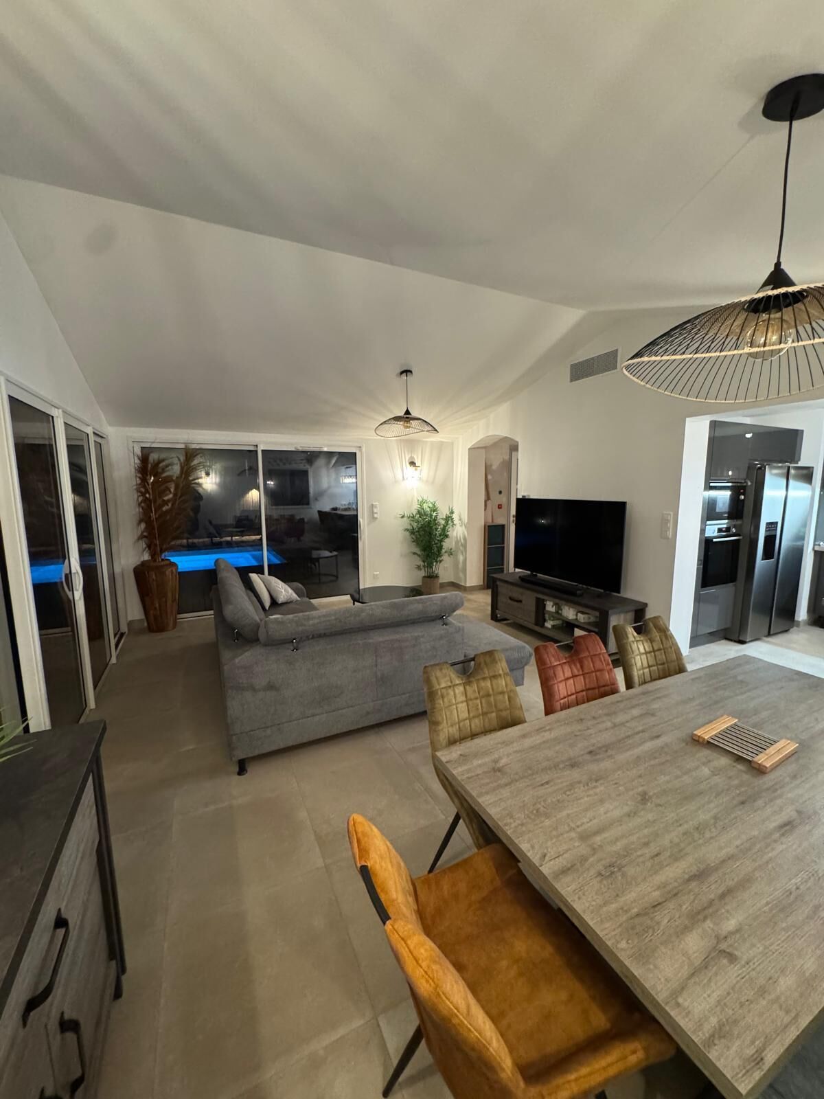 Uma sala de estar moderna com um sofá cinzento, uma mesa de jantar com cadeiras, uma televisão e uma cozinha com frigorífico e forno.