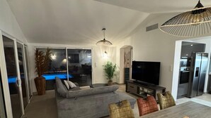 Ein modernes Wohnzimmer mit grauem Sofa, einem Esstisch mit Stühlen, einem Fernseher und einer Küchenzeile mit Kühlschrank und Backofen.