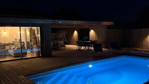 Ein Schwimmbad mit Holzterrasse, einem überdachten Sitzbereich und einem gläsernen Raum mit Sofa und Tisch.