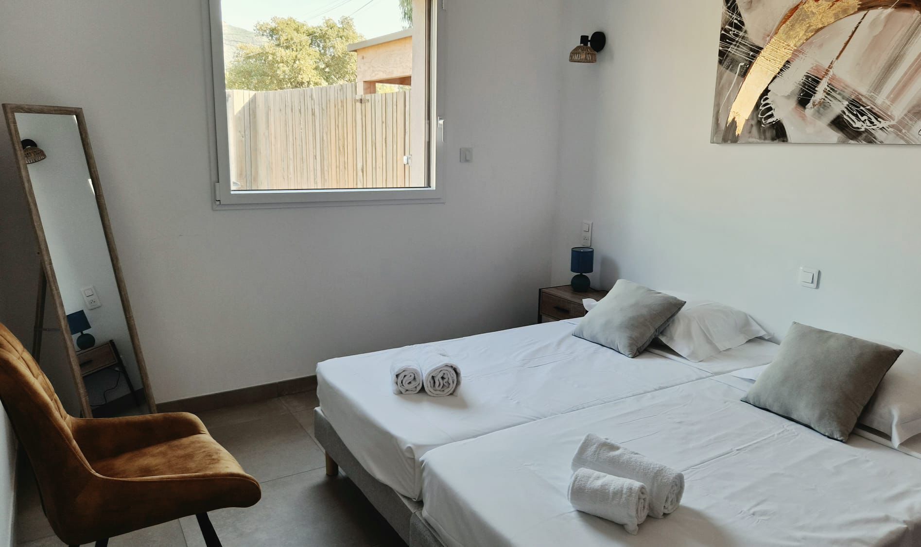 Ein Schlafzimmer mit zwei Betten, einem Sessel, einem Nachttisch mit Lampe, einem Fenster und einem Wandbild.