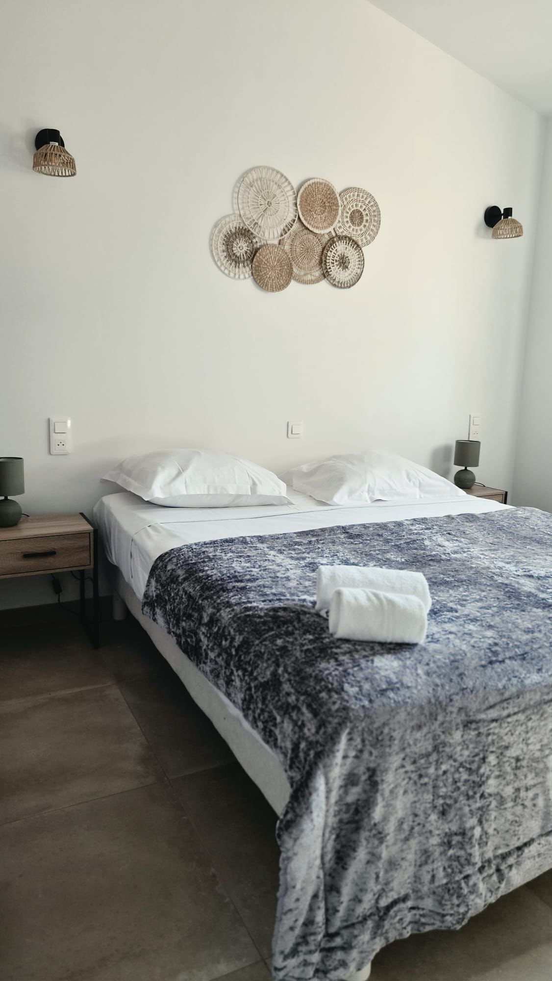Een net opgemaakt bed met een bedsprei met dessin, twee nachtkastjes met lampen, en een wandlamp.