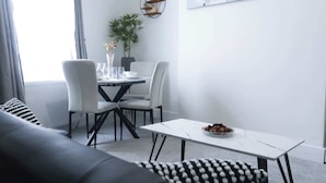 En moderne stue med sofa, spisebord, stole og et lille hvidt bord med en skål frugt.