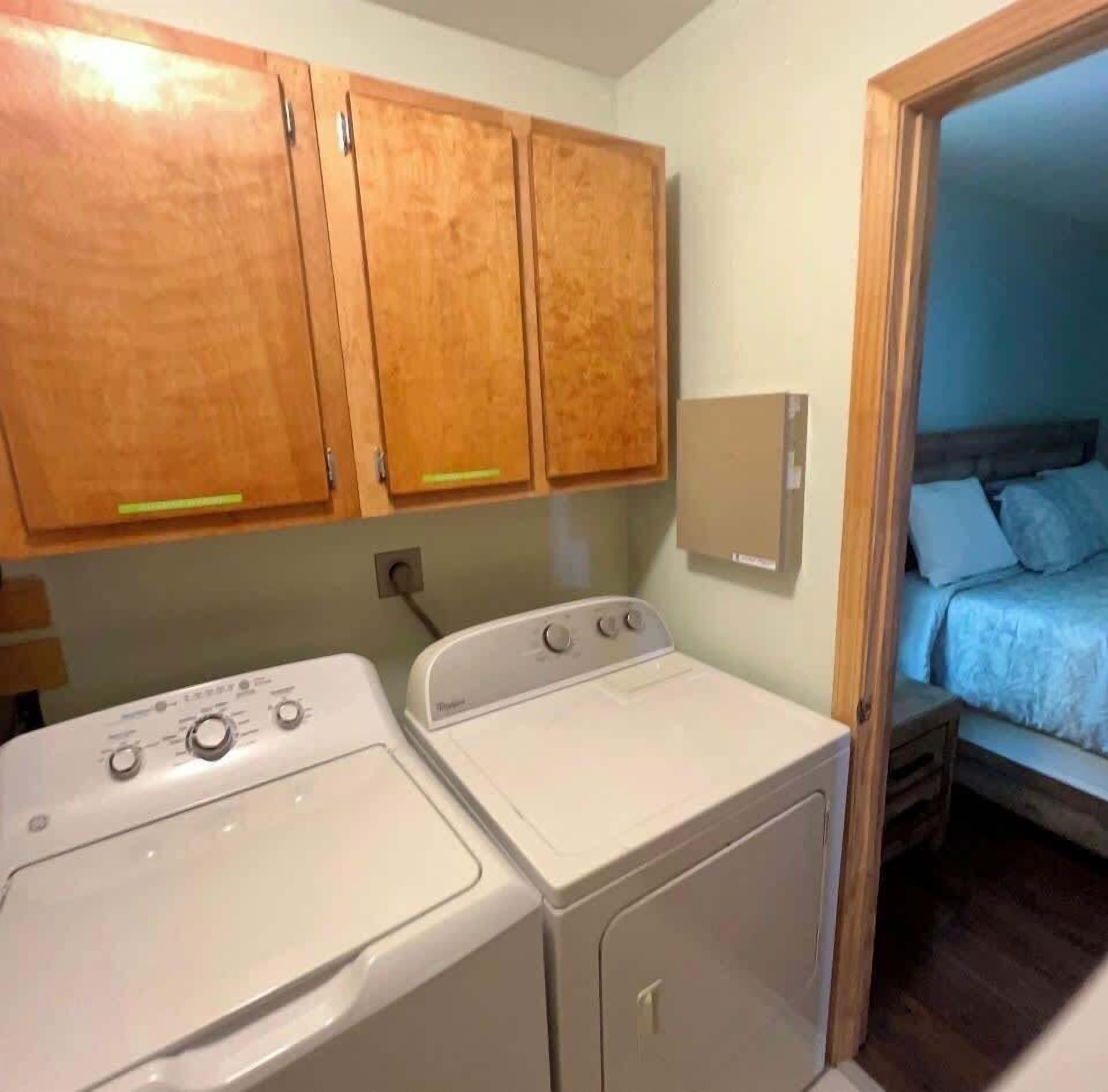 A laundry area with a washer and dryer, wooden cabinets, a small bedside table, and a bed with a headboard.