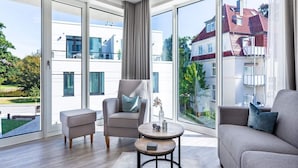 A modern living room with large windows, a grey sofa, a chair, a small table, and a view of a building with red roofs. - Travemunde Beach Retreat (Lübeck)