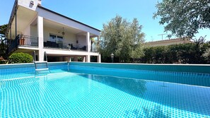 Ein Schwimmbecken mit Fliesen umgeben, ein überdachter Außenbereich mit Balkon und umliegendes Grün.