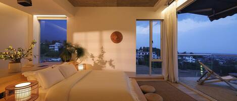 A bedroom with a large bed, a bedside table with a lamp, a window with a view of buildings, and a balcony with a chair.