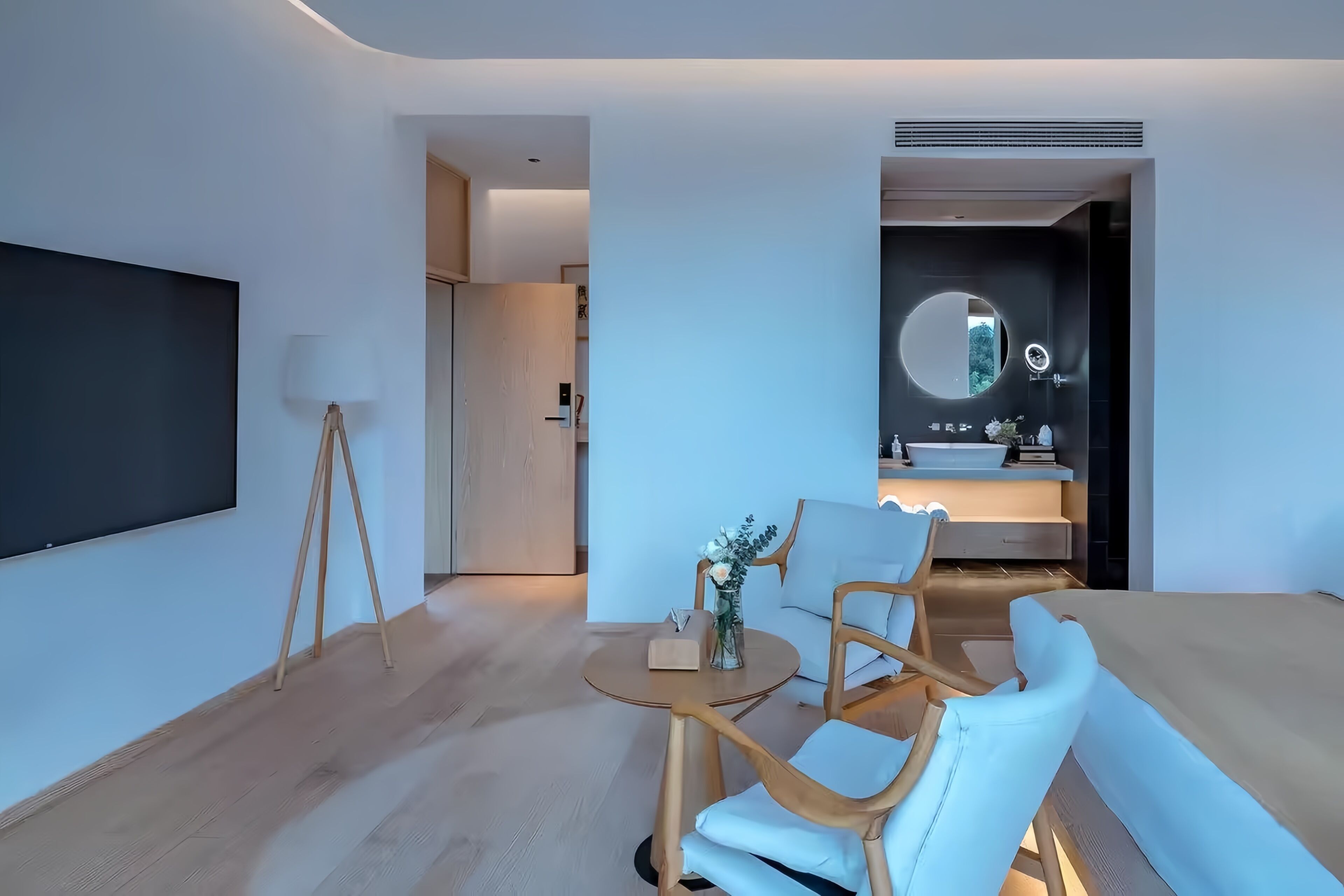 A modern hotel room with a large flat-screen TV, a round mirror above the sink, a sofa, and two wooden chairs.