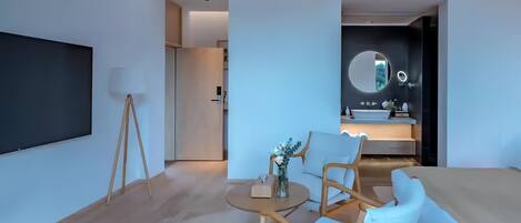 A modern hotel room with a large flat-screen TV, a round mirror above the sink, a sofa, and two wooden chairs.