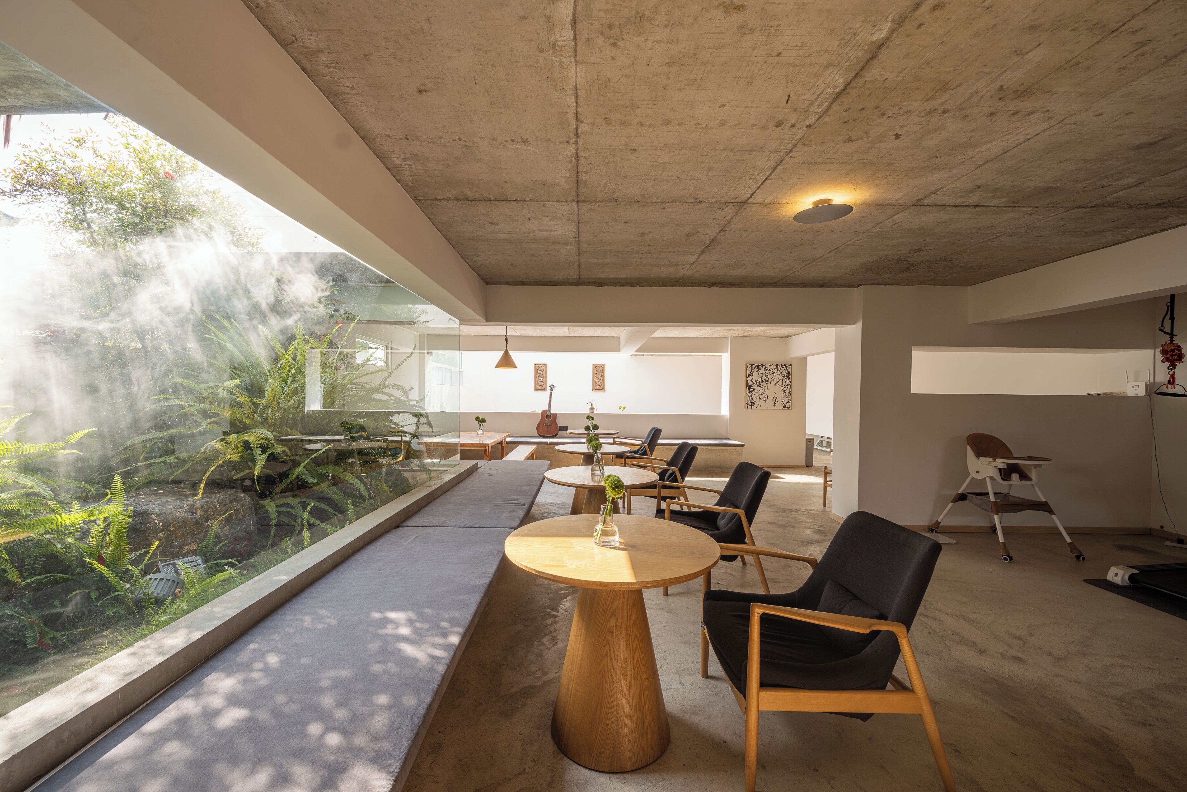 Un intérieur moderne avec un plafond en béton, des tables rondes en bois, des chaises noires et une grande fenêtre donnant sur un jardin.