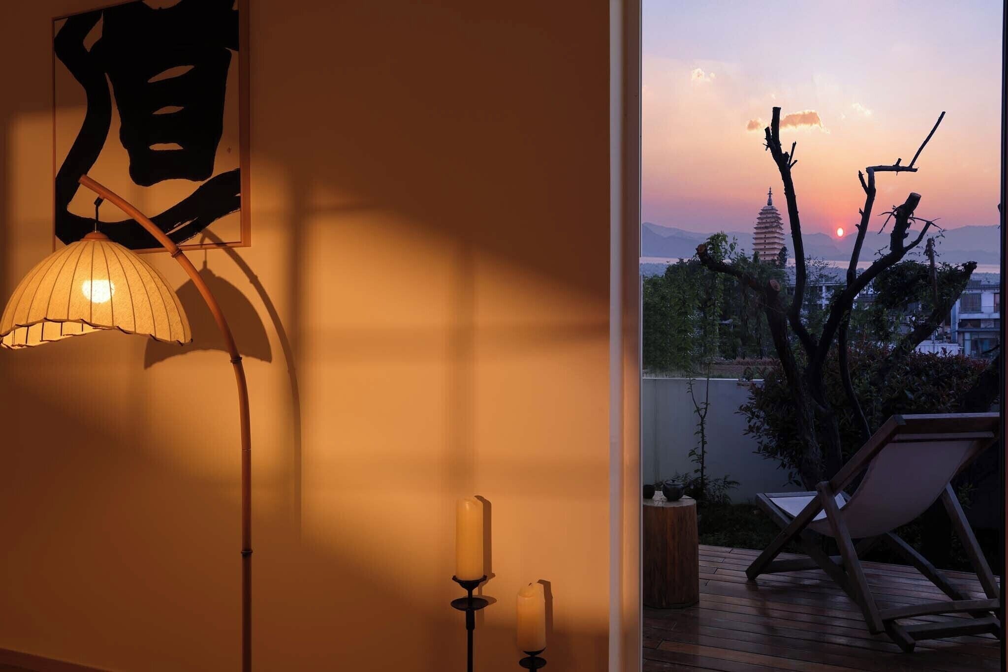 A room with a lamp, candle, and a view of a pagoda at sunset.