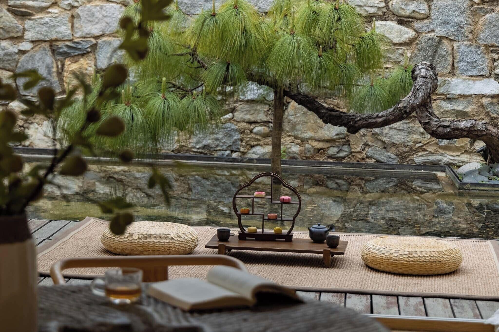 A serene indoor setting with a bonsai tree, a small wooden stand with colorful objects, and a table with a woven mat.