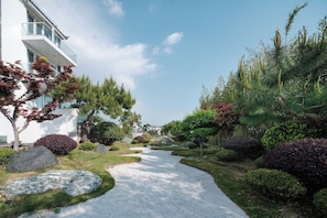Un chemin mène à un bâtiment moderne doté d’un balcon, lequel est entouré de jardins aménagés avec différents arbustes et arbres.
