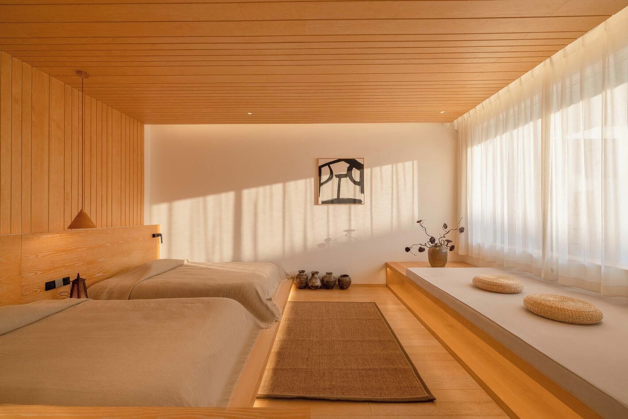 A minimalist bedroom with two beds, a wooden ceiling, and a large window with sheer curtains.