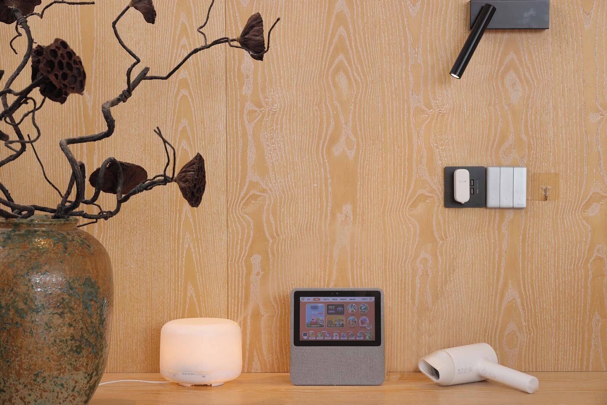 Una pared de madera con una planta decorativa, una vela blanca, una tableta y un secador de pelo sobre una superficie de madera.