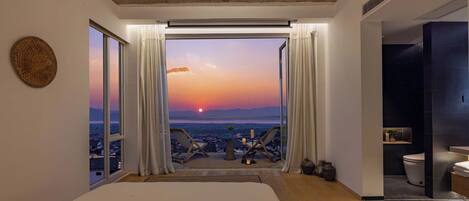 A bedroom with a large bed, a view of the sunset, and a balcony with chairs.