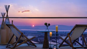 Une terrasse avec deux chaises en bois, une petite table avec des bougies et une vue sur un coucher de soleil se reflétant sur une étendue d’eau et des montagnes.