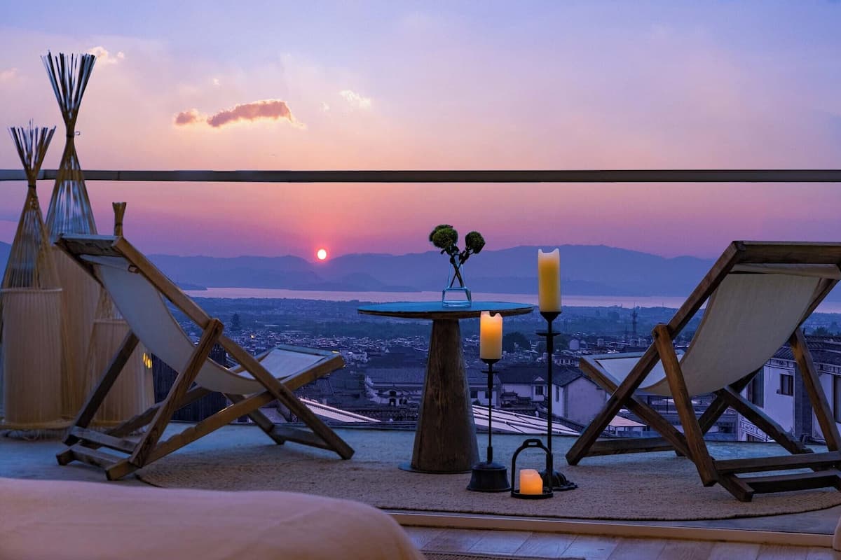 A terrace with two wooden chairs, a small table with candles, and a view of a sunset over a body of water and mountains.