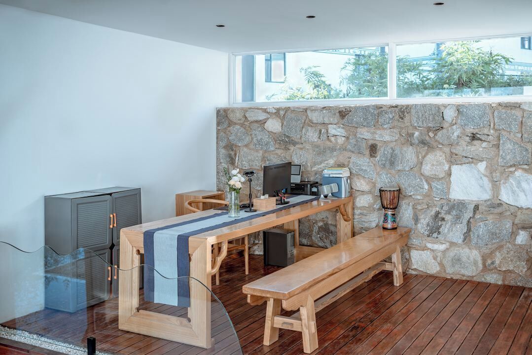 Une chambre moderne avec un mur en pierre, une table en bois, un banc, un téléviseur et une fenêtre donnant sur un paysage verdoyant.