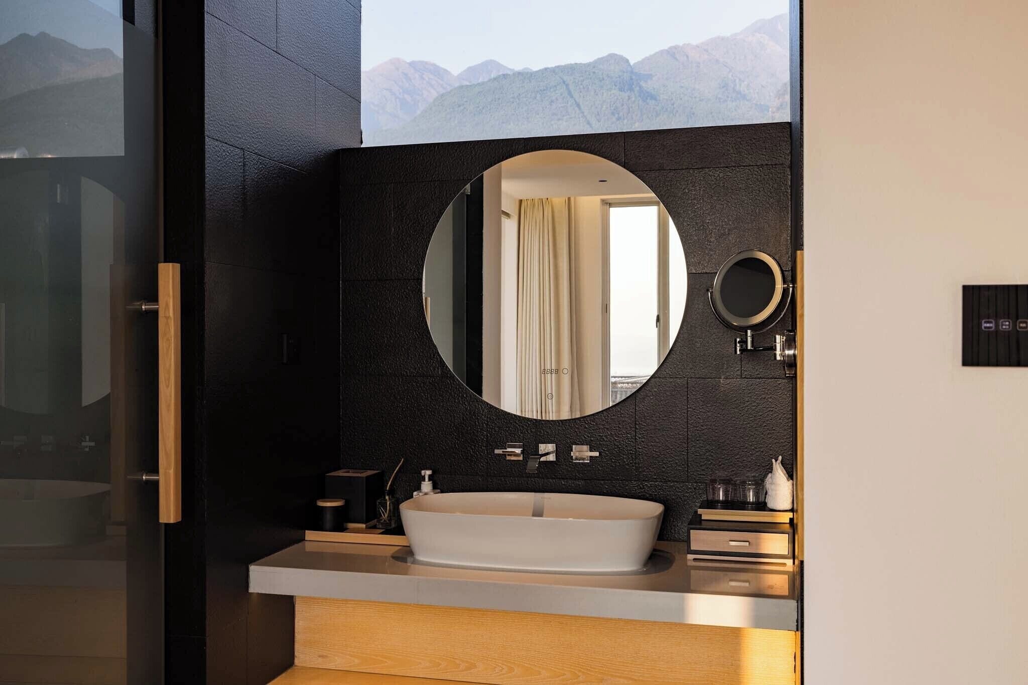 Een moderne badkamer met een grote ovale spiegel, een vrijstaand wastafelmeubel en uitzicht op bergen door een glazen deur.