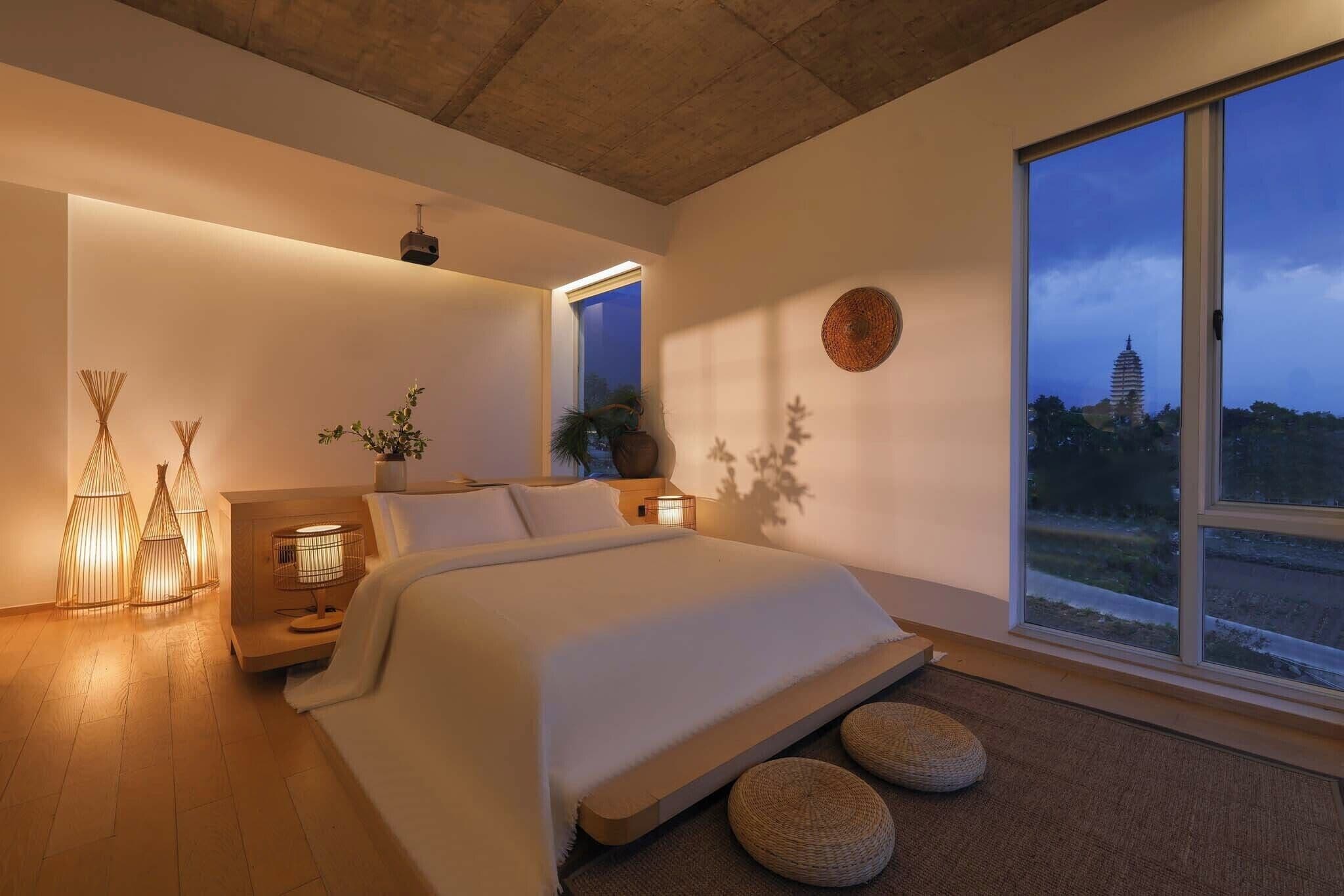 Une chambre moderne avec une grande fenêtre, un lit, des lampes de chevet et une vue sur une tour distante.