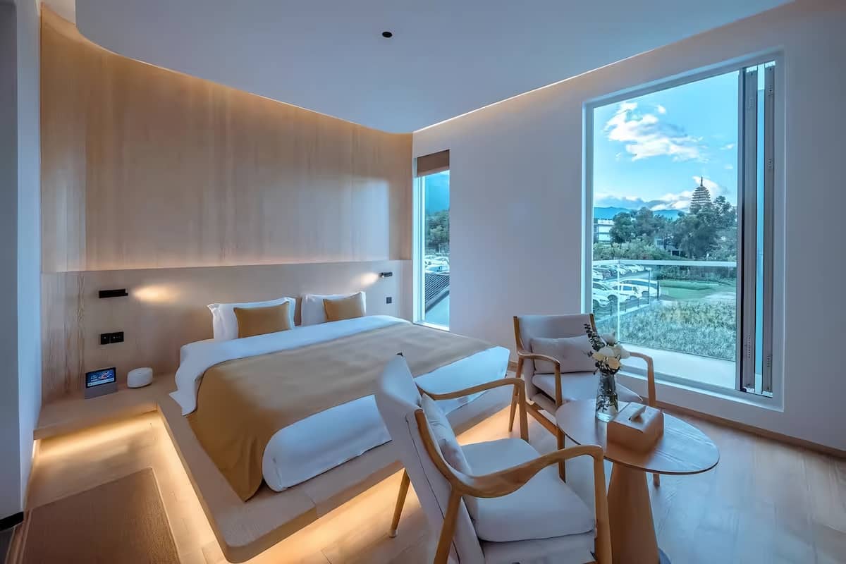 A modern hotel room with a large bed, a small table with chairs, and a view of a cityscape through a large window.
