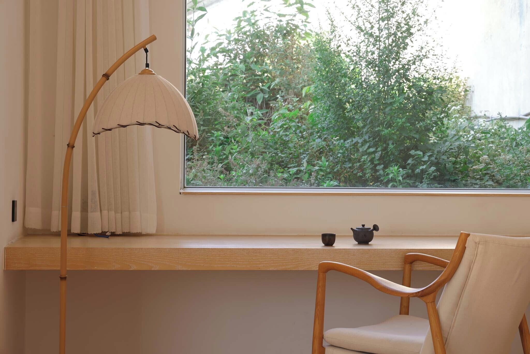 Een kamer met een houten bureau, een stoel, een vloerlamp en een raam met uitzicht op groen.