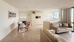 A modern living room with a sofa, a dining table with chairs, and a kitchen area with cabinets and a microwave. - Allamanda Place Unit 11 Bulcock Beach Waterfront Views (Caloundra)