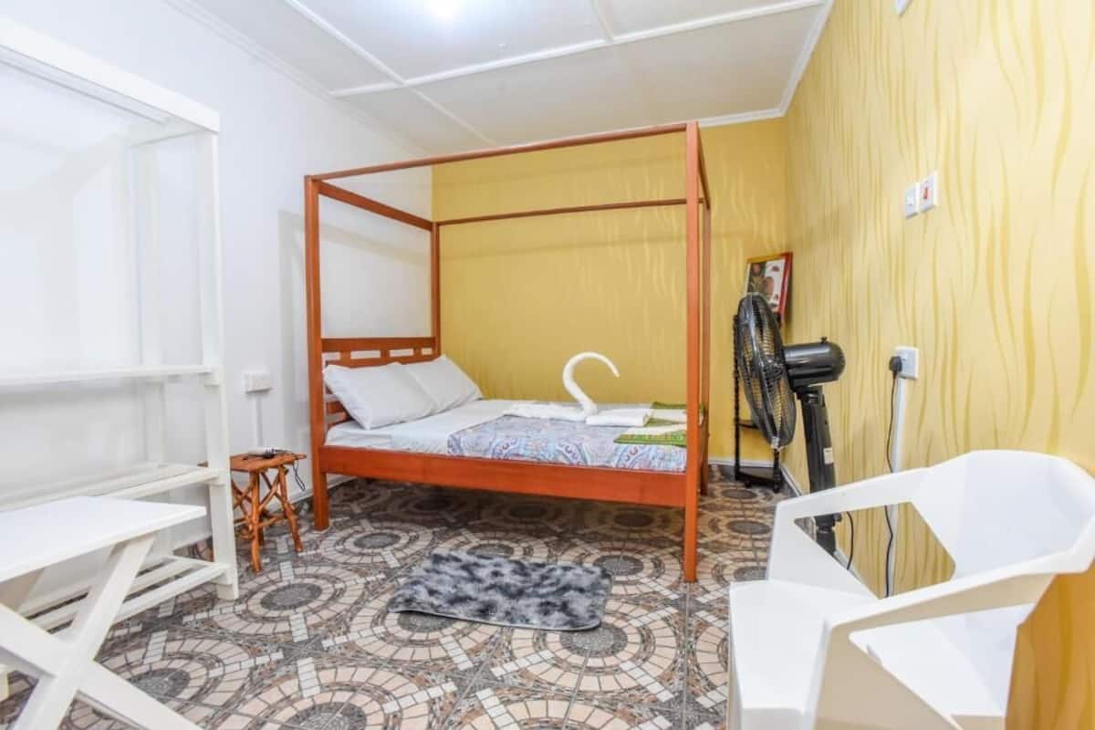 Una cama individual con estructura de madera, una silla blanca, una mesita pequeña, un ventilador y un suelo con diseño.