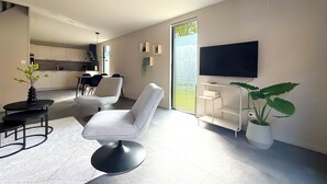 Ein modernes Wohnzimmer mit Flachbildfernseher, einem Essbereich mit Stühlen, einer Küche im Hintergrund und einem großen Fenster.
