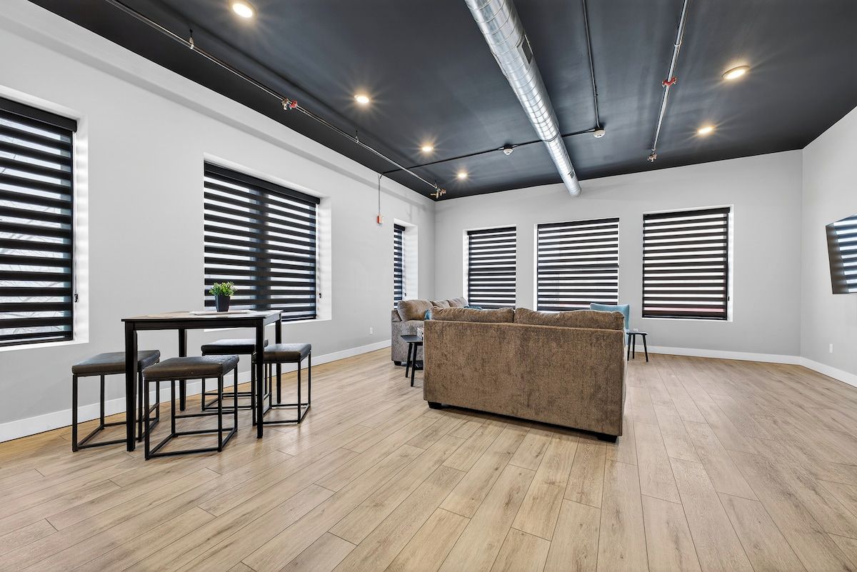 A spacious living area with a dark ceiling, large windows with blinds, a brown sofa, and a dining table with chairs.