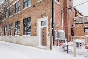Un bâtiment en briques avec le numéro 2 sur la porte, une enseigne indiquant « LOFTS », et des tables et des chaises d’extérieur recouvertes de neige.